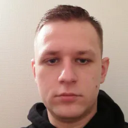 Portret młodego mężczyzny w czarnej bluzie z kapturem na tle jasnej, teksturowanej ściany.