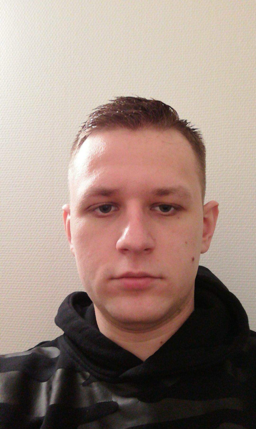 Portret młodego mężczyzny w czarnej bluzie z kapturem na tle jasnej, teksturowanej ściany.