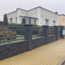 Nowoczesne ogrodzenie z szarego kamienia łupanego i poziomych, metalowych przęseł w kolorze grafitowym, na tle domu z numerem 33, widok z chodnika wyłożonego kostką.