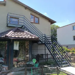 Zewnętrzne schody metalowe z balustradą prowadzące na balkon, zamontowane przy dwukondygnacyjnym budynku mieszkalnym z drewnianą altaną w ogrodzie.