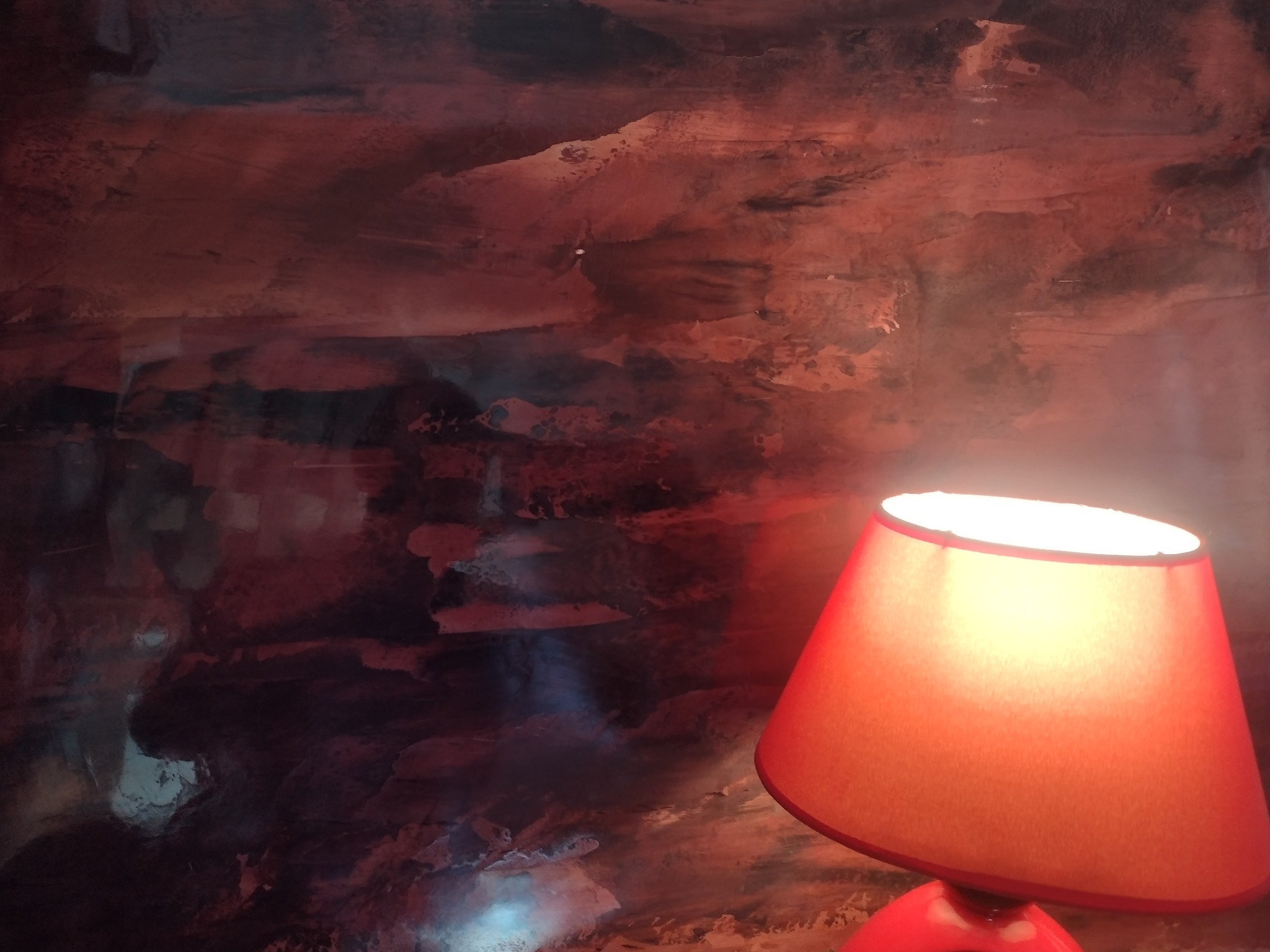 Czerwona lampka nocna z abażurem na tle ściany z dekoracyjnym malowaniem w odcieniach czerwieni i szarości.