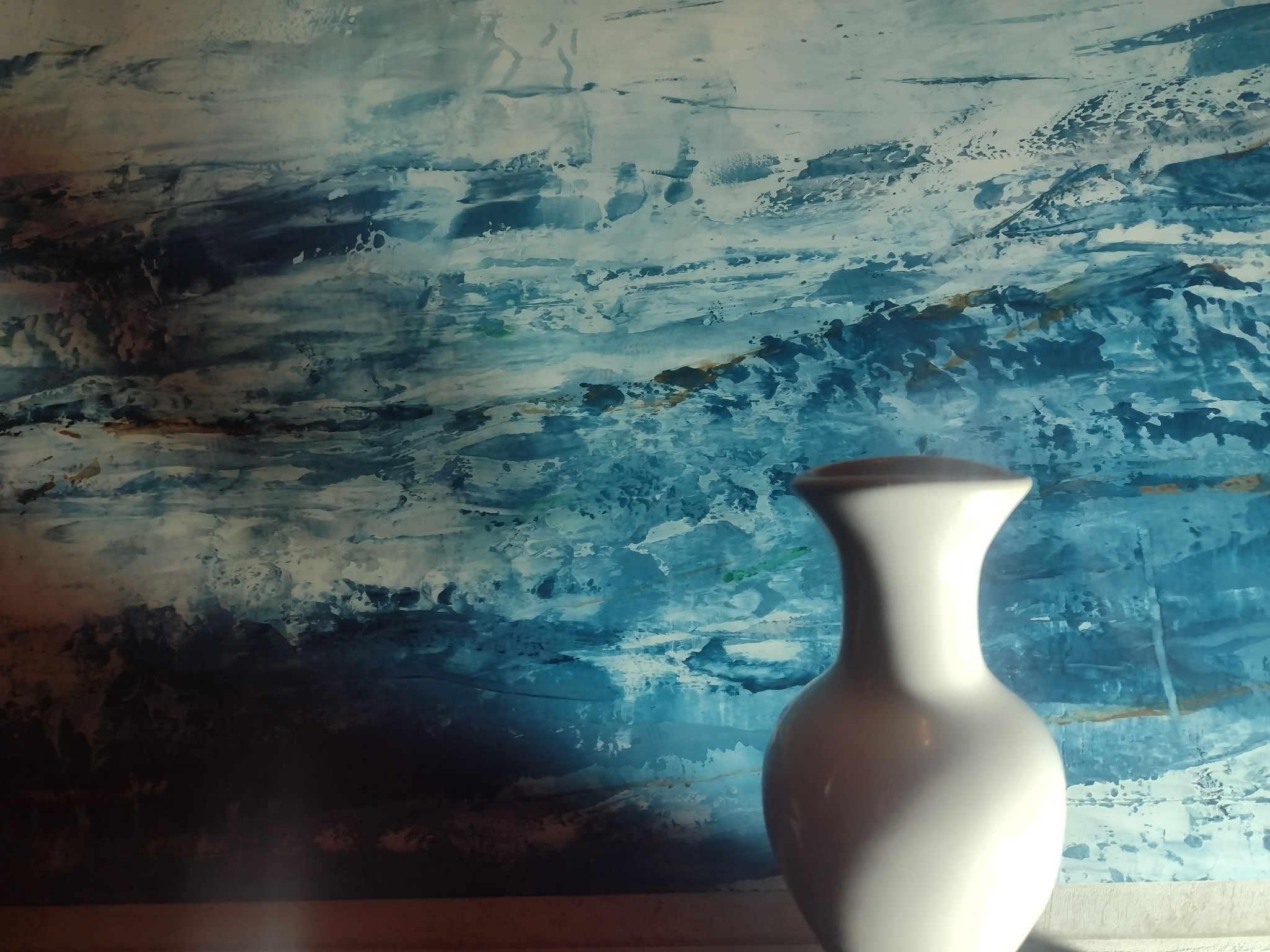 Biały wazon na tle ściany z abstrakcyjnym malowaniem w odcieniach błękitu i bieli, przypominającym wzburzone morze.