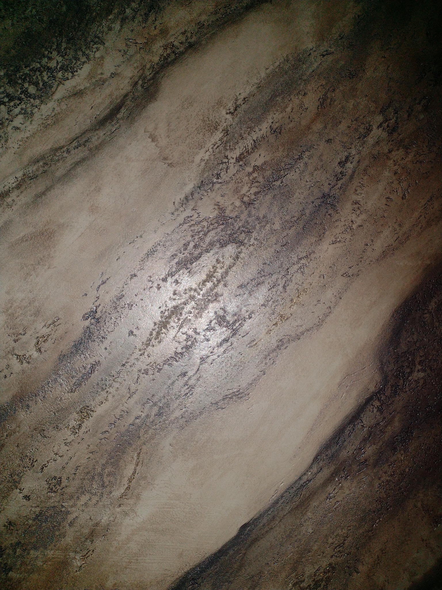 Beżowa ściana z dekoracyjną fakturą imitującą marmur, z ciemnymi żyłkami i delikatnym połyskiem, oświetlona z góry.