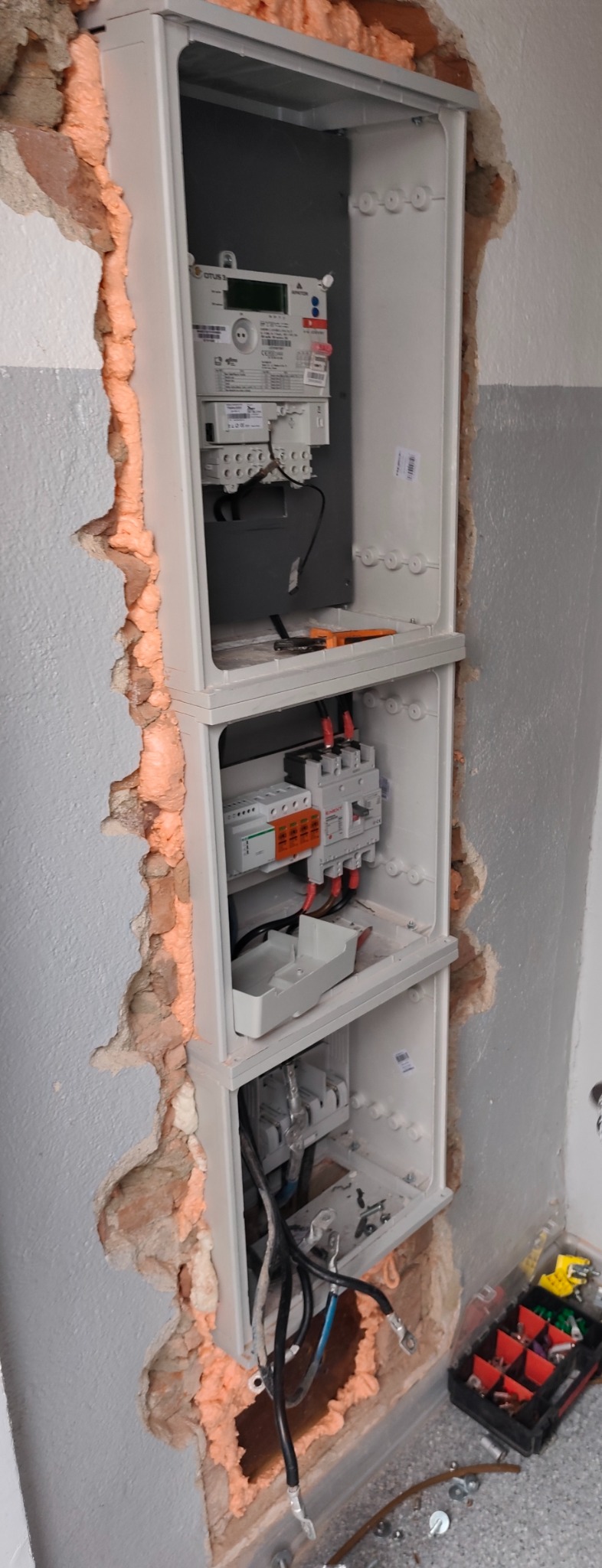 Rozdzielnica elektryczna w trakcie montażu, widoczne zabezpieczenia, licznik i przewody. Ściana wokół obudowy wypełniona pianką montażową. Skrzynka z narzędziami na podłodze.