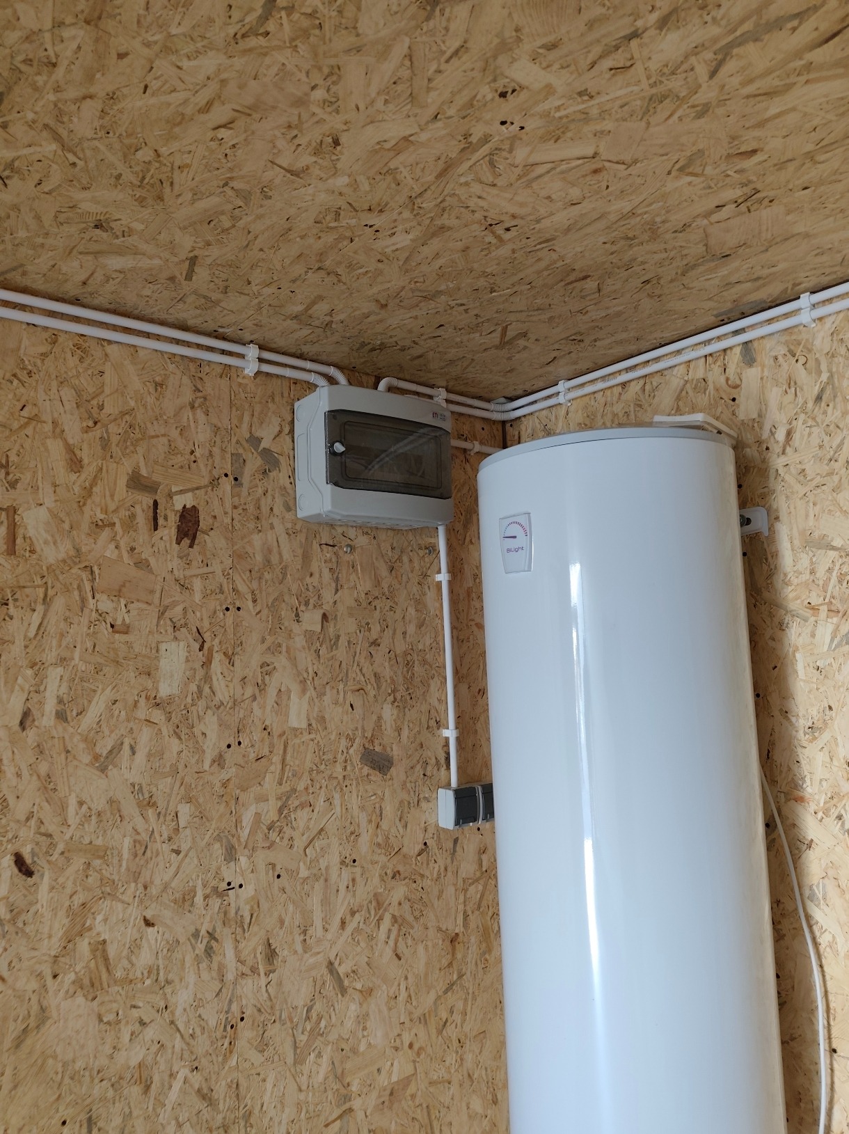 Biały bojler elektryczny zamontowany na ścianie z płyty OSB, obok szara skrzynka elektryczna z przezroczystymi drzwiczkami i białe kable w peszlu.