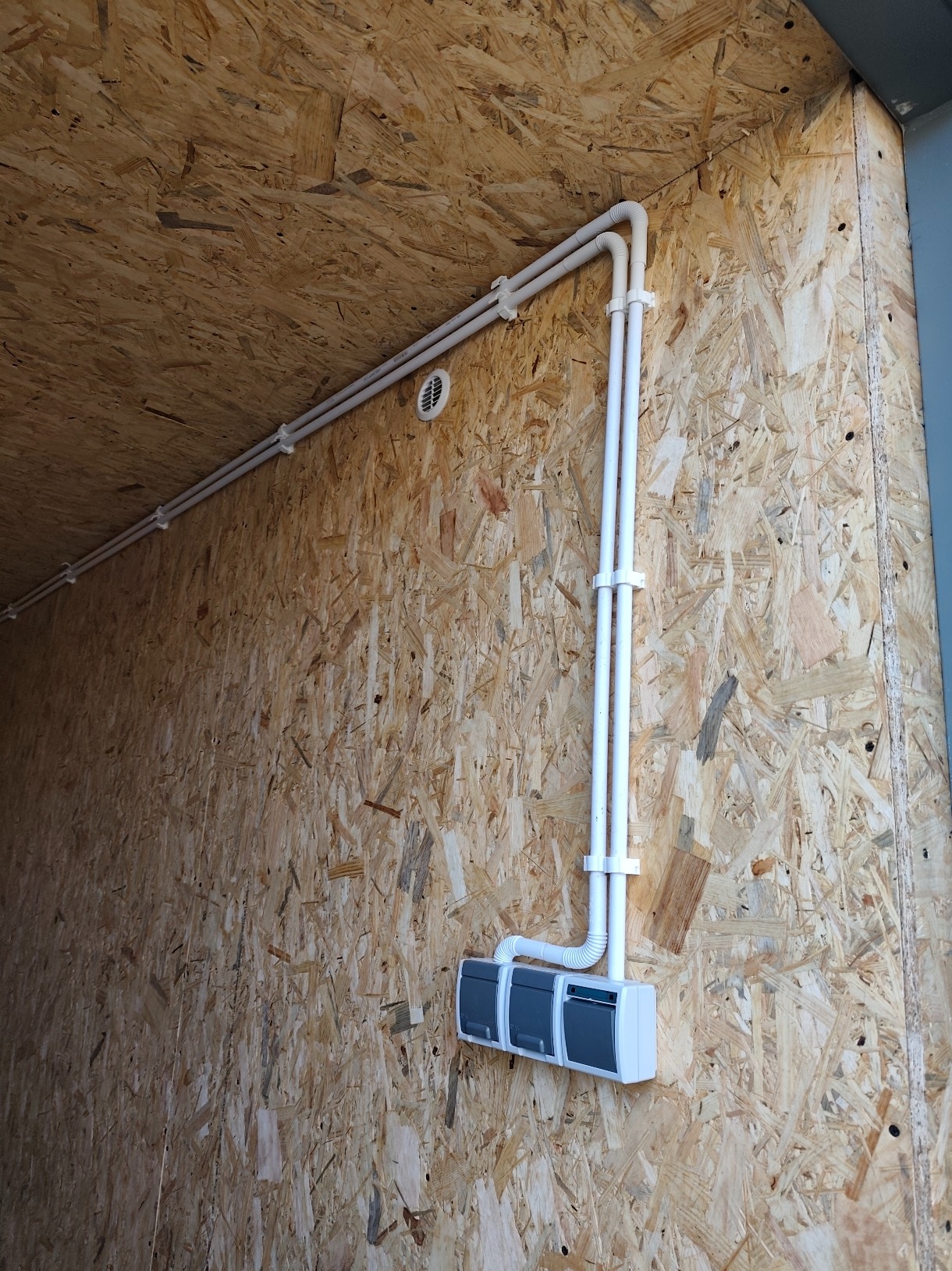 Białe rury instalacji elektrycznej przymocowane do ściany z płyty OSB. Widoczne gniazdka i wentylacja. Ujęcie z dołu, podkreślające pionowy przebieg instalacji.