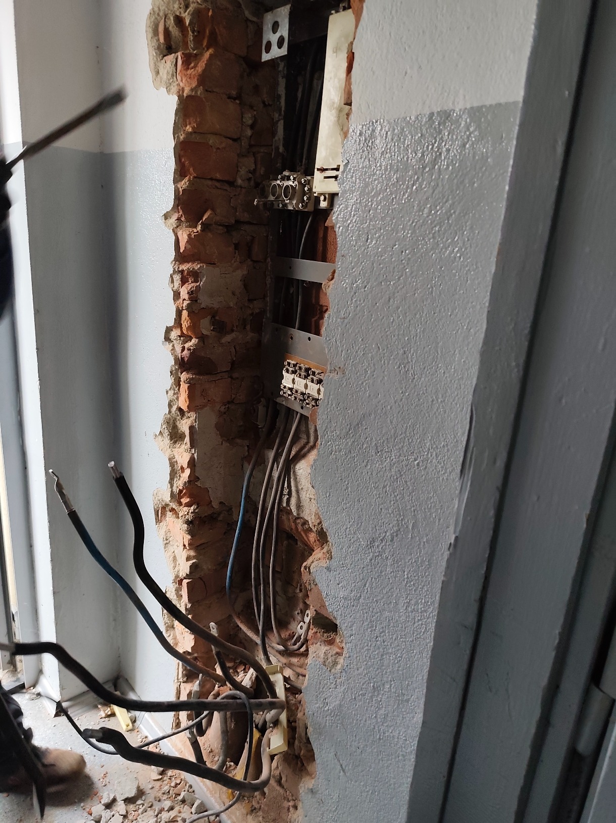 Odsłonięta instalacja elektryczna w ścianie z cegły, widoczne przewody i puszki przyłączeniowe. Fragment ściany pomalowany na szaro. Widoczne uszkodzenia ściany.