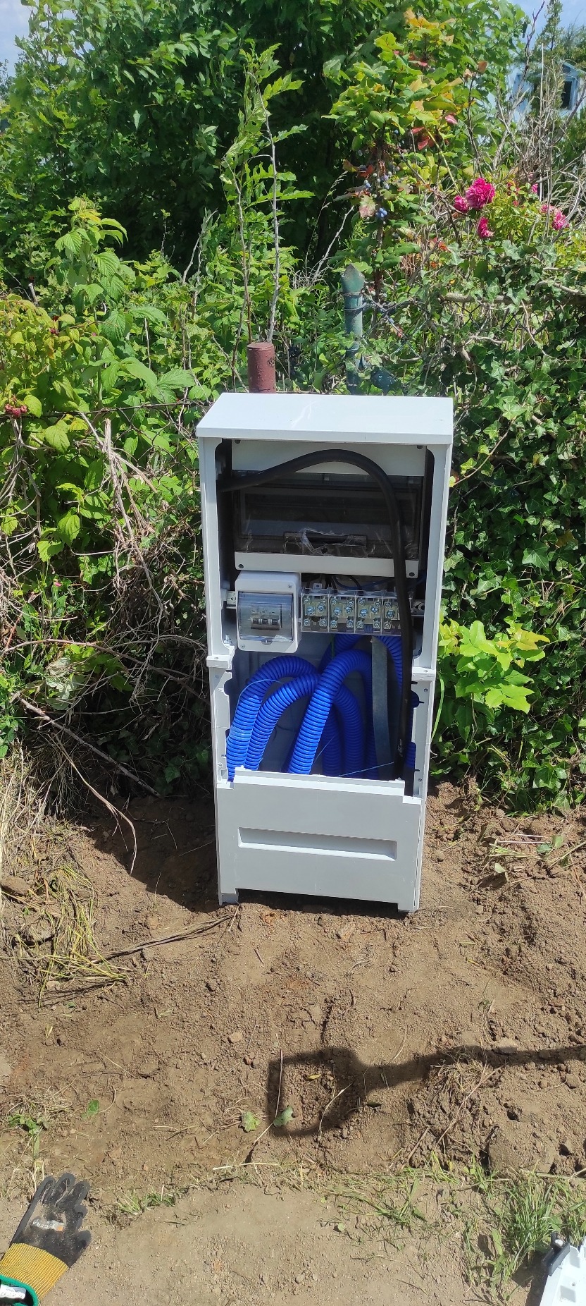 Otwarta, biała skrzynka elektryczna z niebieskimi rurami karbowanymi i przewodami, umieszczona na ziemi, otoczona zielenią. Widoczne zabezpieczenia i elementy instalacji.
