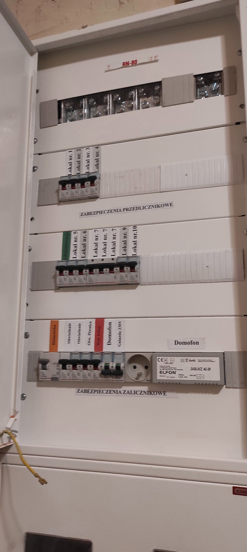 Rozdzielnica elektryczna z bezpiecznikami oznaczonymi dla różnych obwodów (lokale, oświetlenie, domofon), zasilacz AC-20 Elfon i gniazdo 230V. Widok z bliska.