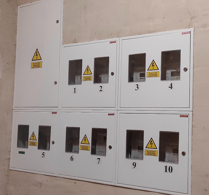 Rozdzielnia elektryczna z numerowanymi licznikami w białych obudowach, ostrzeżenie o napięciu. Widok z bliska na instalację elektryczną w budynku.