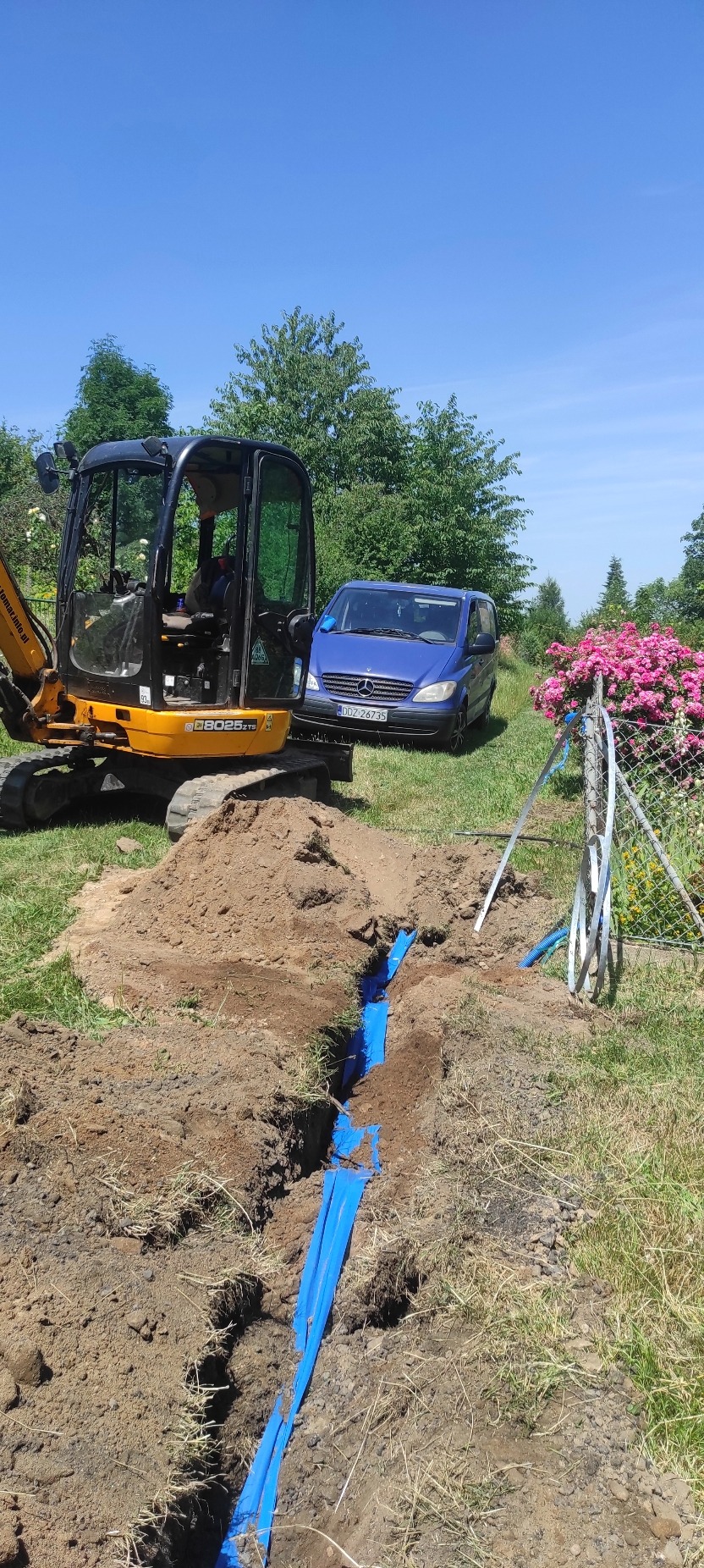 Niebieskie rury w wykopie, zabezpieczone folią, z koparką i autem w tle. Prace ziemne w ogrodzie, słoneczny dzień.