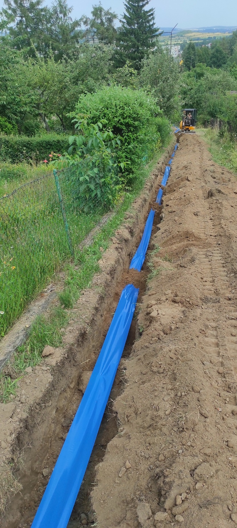 Wykopany rów z niebieską rurą ochronną, prawdopodobnie pod kable elektryczne, w tle koparka i zieleń. Prace ziemne w toku.