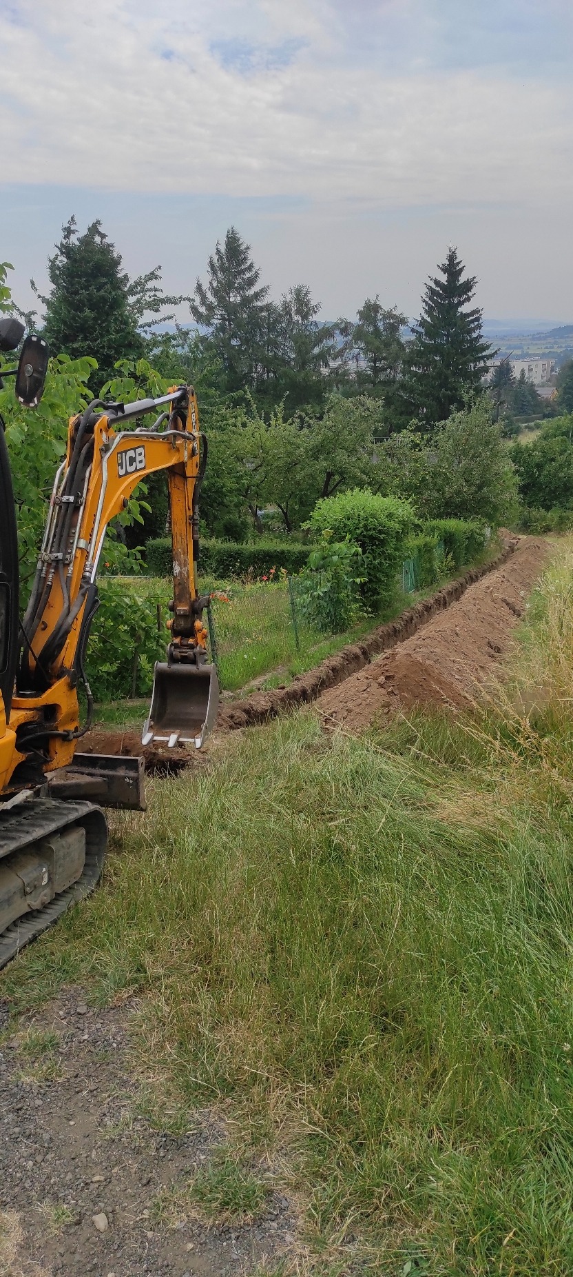 Żółta koparka JCB wykopuje wąski rów w ziemi na tle zieleni i drzew, z widokiem na miasto w oddali. Prace ziemne w toku, widoczne ślady świeżo wykopanej ziemi.