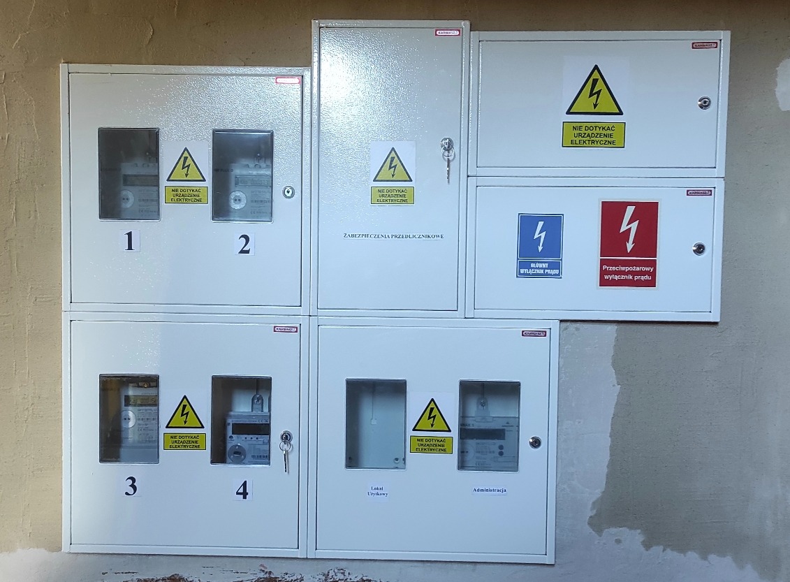 Zestaw białych skrzynek elektrycznych z licznikami i ostrzeżeniami, zamontowanych na ścianie. Widoczne numery identyfikacyjne i oznaczenia bezpieczeństwa.