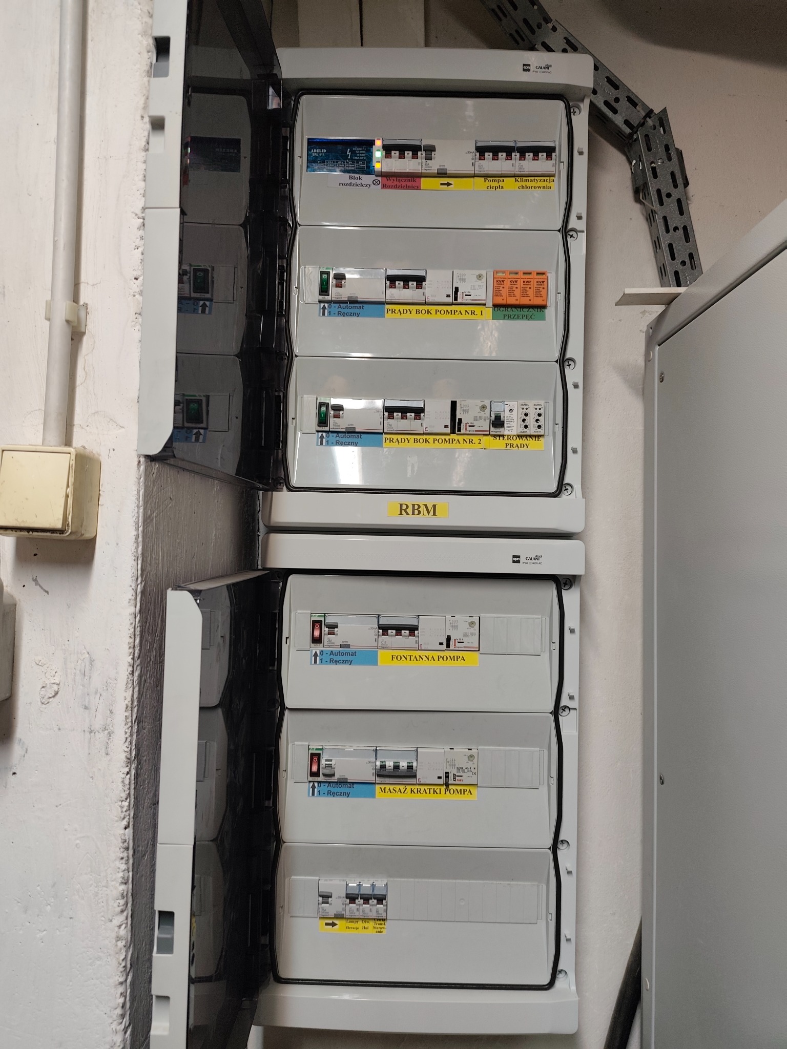 Rozdzielnia elektryczna z opisami obwodów w Bielawie. Widoczne zabezpieczenia i wyłączniki. Instalacja w szarej obudowie, zamontowana na ścianie.