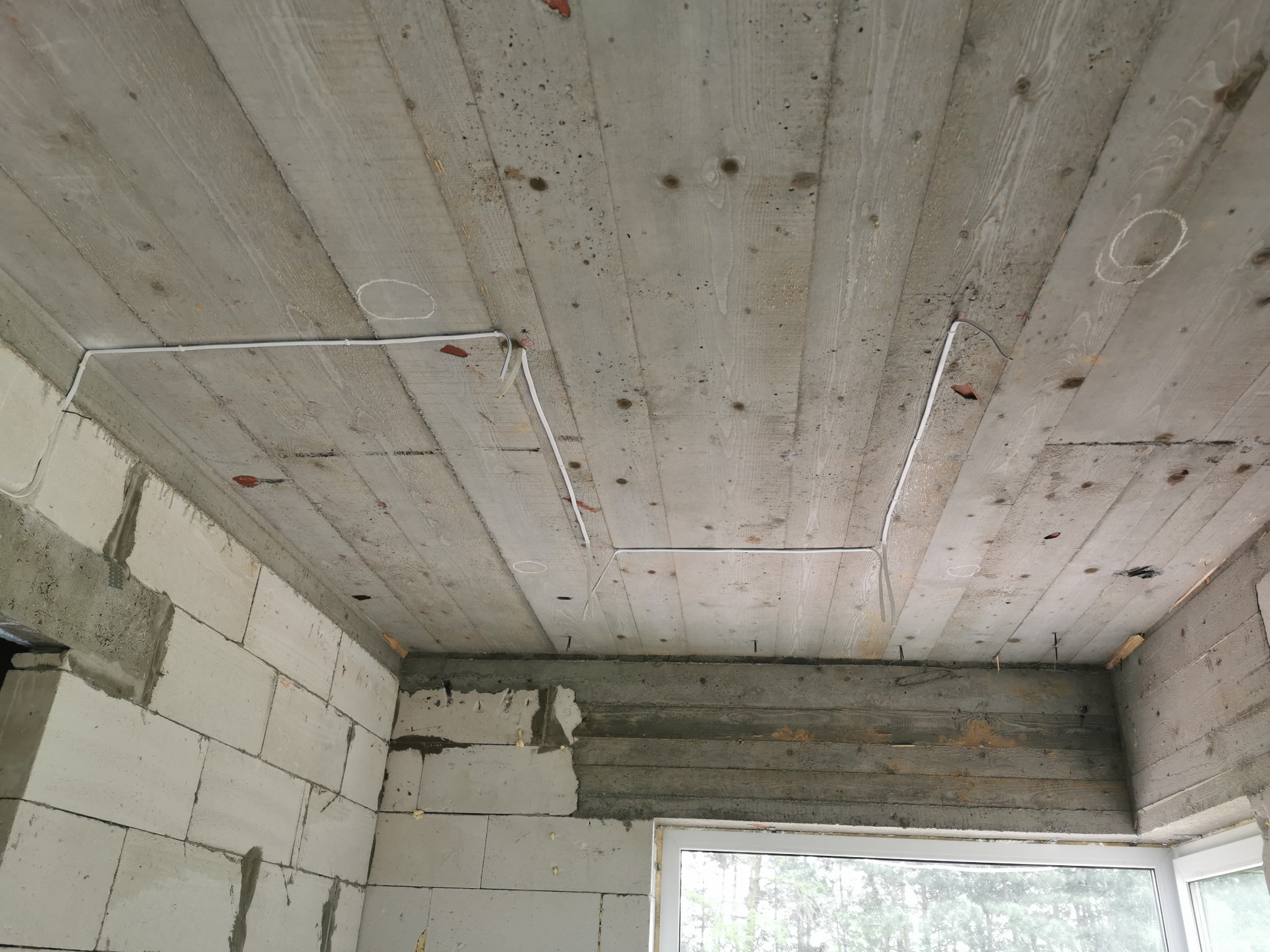 Surowy betonowy strop z widocznymi deskami szalunkowymi i prowizorycznie ułożonymi przewodami elektrycznymi w białych listwach, ściany z bloczków betonowych w stanie surowym.