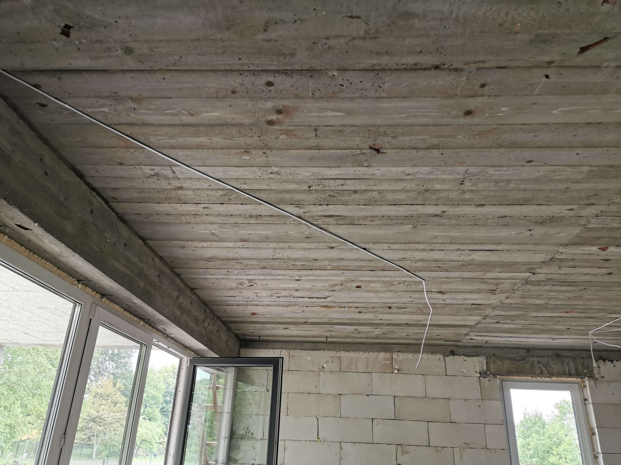 Surowy betonowy strop z widocznymi śladami szalunku i poprowadzoną białą instalacją elektryczną w rurce, okna w stanie surowym otwarte na zewnątrz, ściany z bloczków betonowych.