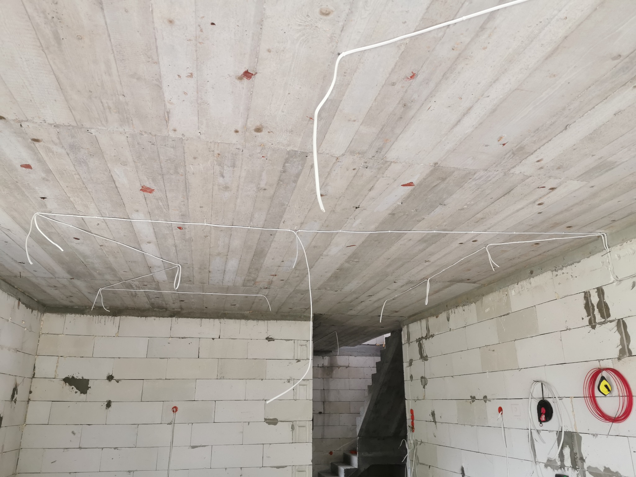 Surowy strop betonowy z widocznymi śladami szalunków i przewodami elektrycznymi w białej izolacji, wystającymi ze ściany z betonu komórkowego. Widoczny fragment klatki schodowej w tle.