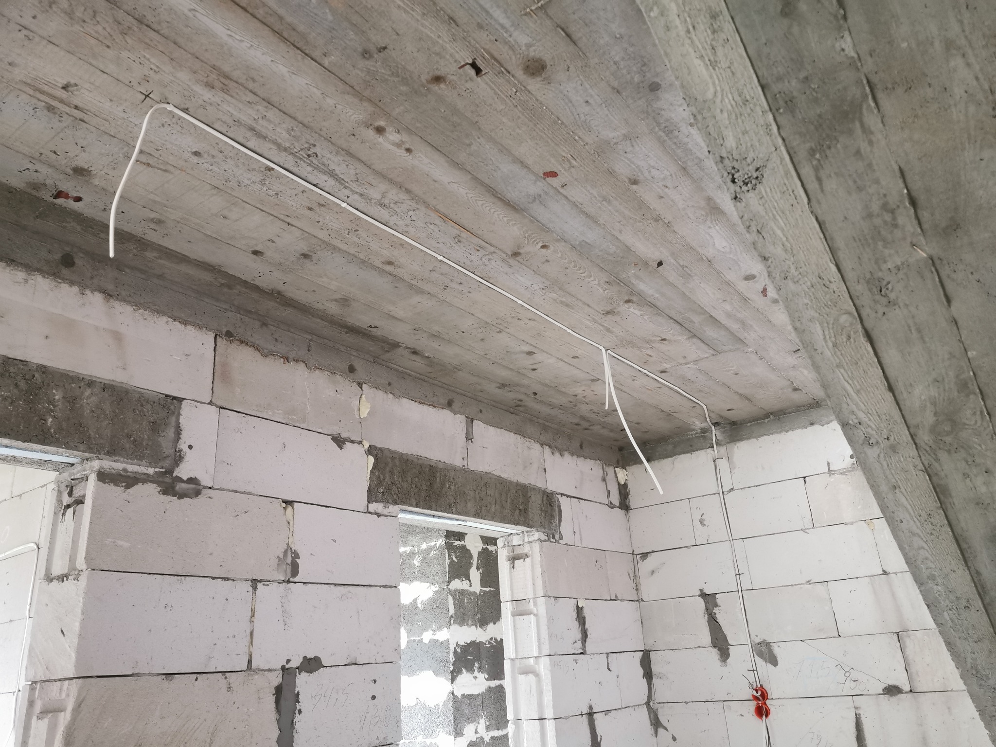 Biały przewód elektryczny prowadzony po betonowym suficie w surowym pomieszczeniu z białych bloczków, widoczne mocowania przewodu i fragment czerwonej puszki elektrycznej na ścianie.