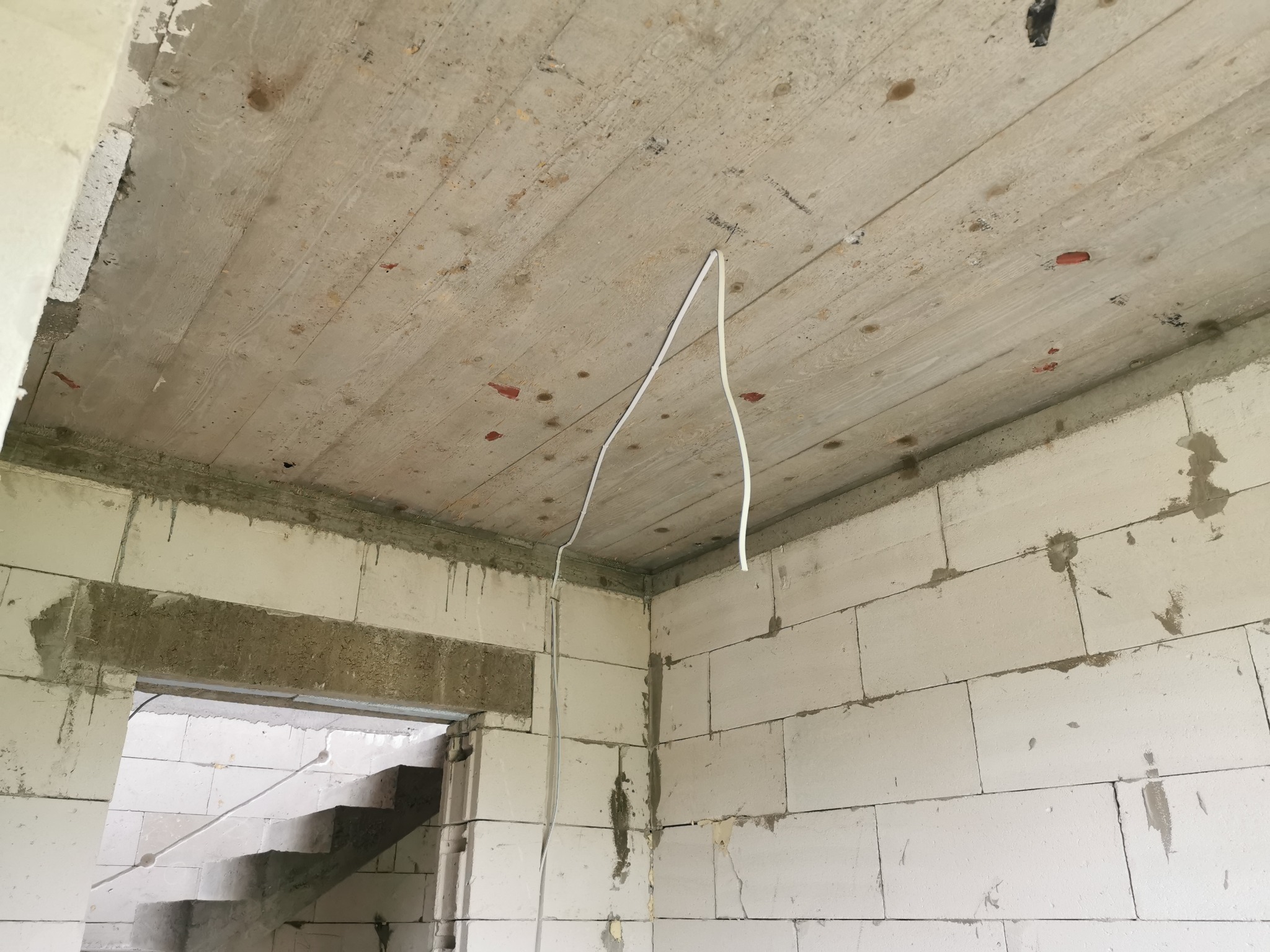 Surowy stan wnętrza w trakcie budowy z widocznymi przewodami elektrycznymi zwisającymi z sufitu, ściany z białych bloczków betonowych, widoczne ślady zaprawy.