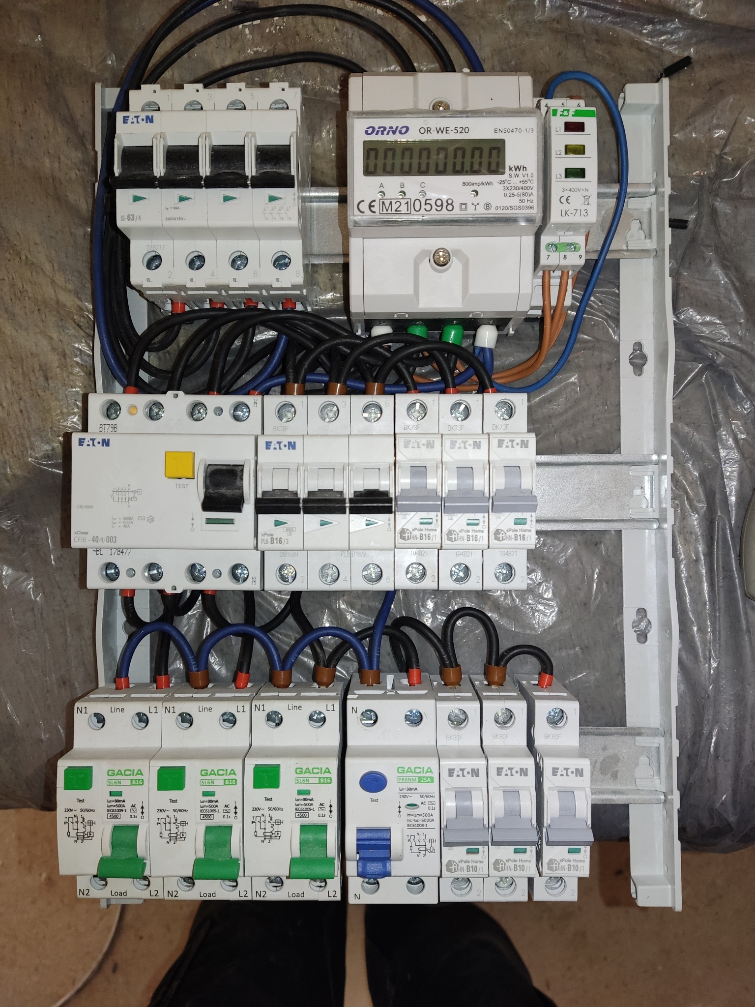 Rozdzielnica elektryczna z zainstalowanymi bezpiecznikami Eaton, Gacia i licznikiem Orno, widoczne przewody elektryczne, montaż w skrzynce na ścianie pokrytej folią