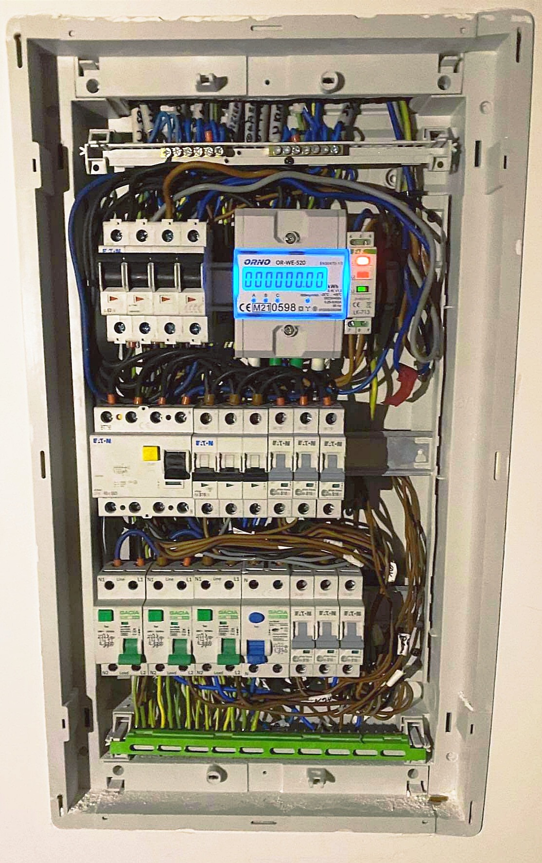 Otwarta skrzynka rozdzielcza z licznikiem ORNO OR-WE-520 z podświetlanym wyświetlaczem, bezpiecznikami EATON i GACIA, widoczne okablowanie w różnych kolorach.