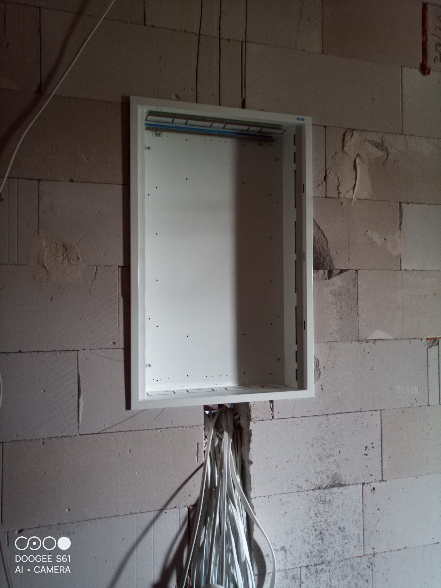 Pusta, biała skrzynka rozdzielcza zamontowana w surowej ścianie z betonu komórkowego, z wystającymi kablami elektrycznymi u dołu.