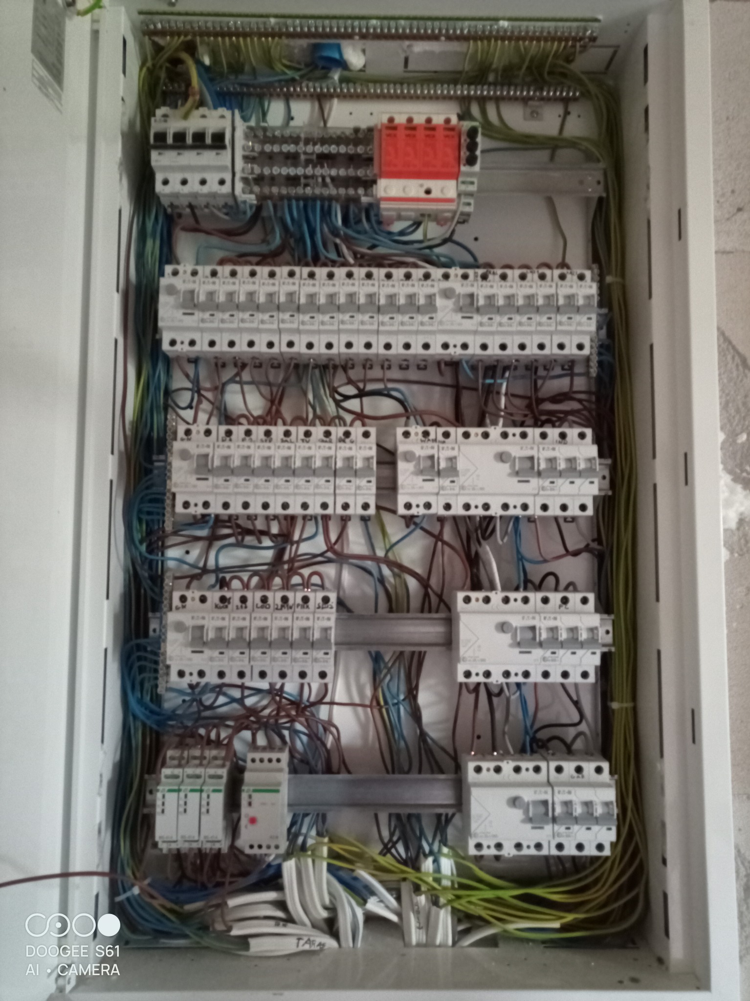 Otwarta skrzynka rozdzielcza z licznymi bezpiecznikami, przewodami w różnych kolorach i listwami zaciskowymi, widoczne ślady po montażu na ścianie.