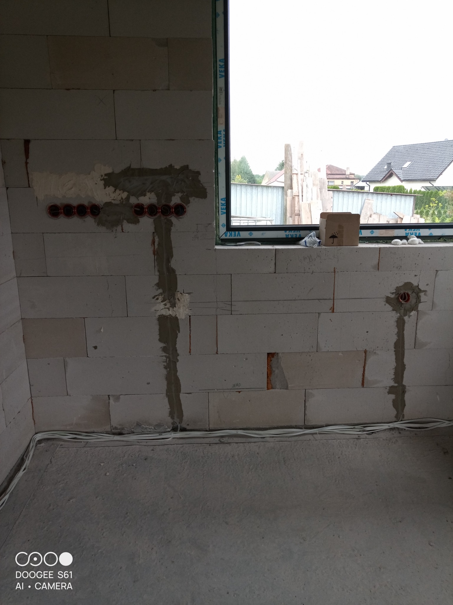 Surowy stan wnętrza w trakcie prac: widoczne puszki pod gniazdka elektryczne w ścianie z betonu komórkowego, częściowo zamaskowane gładzią, oraz przewody elektryczne ułożone wzdłuż podłogi, z oknem...