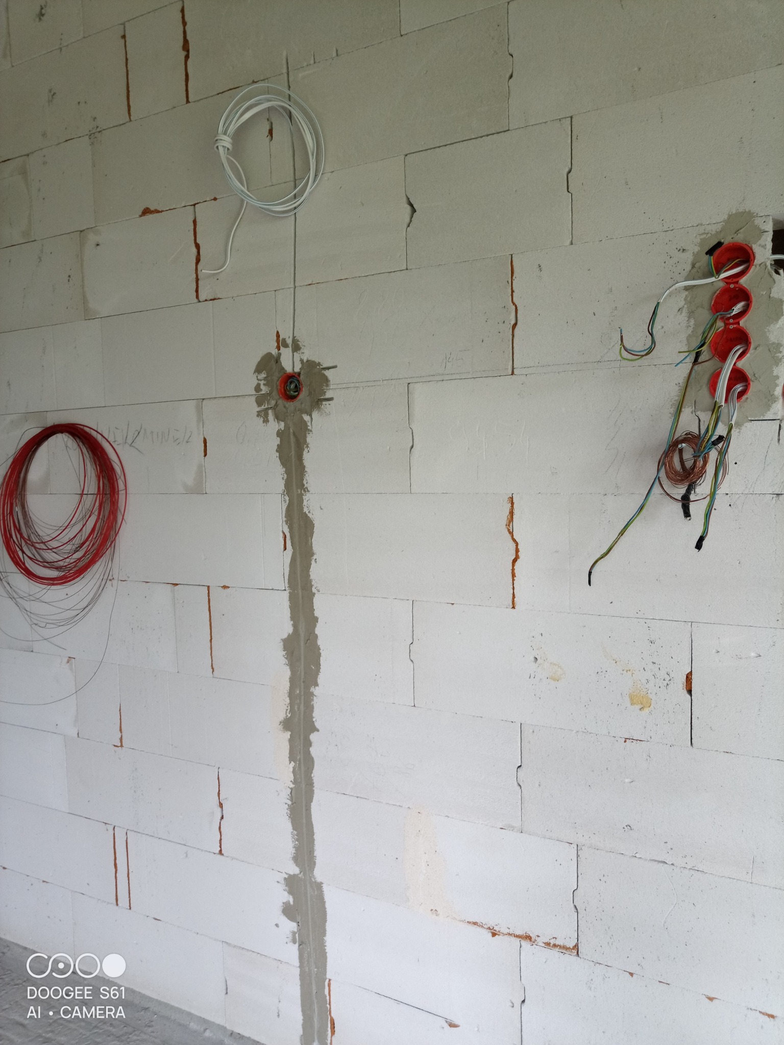 Ściana z betonu komórkowego w trakcie prac elektrycznych: widoczne kable w kolorach białym i czerwonym, puszki instalacyjne w kolorze pomarańczowym oraz bruzda zalana gipsem.