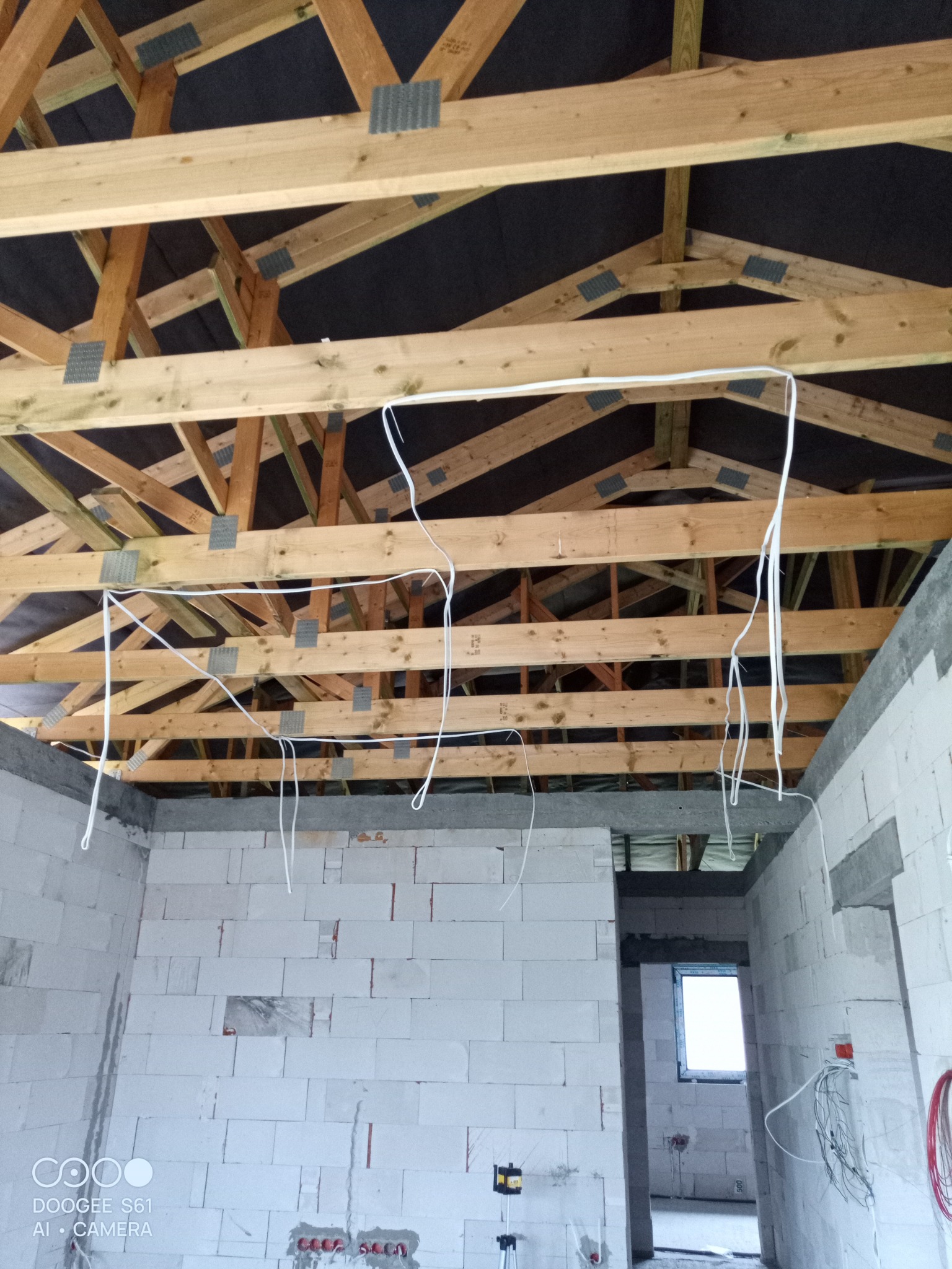 Surowy stan wnętrza domu w budowie, widoczne drewniane krokwie dachu z metalowymi łącznikami, wstępnie poprowadzone białe kable elektryczne zwisające z sufitu, ściany z białych bloczków betonowych...