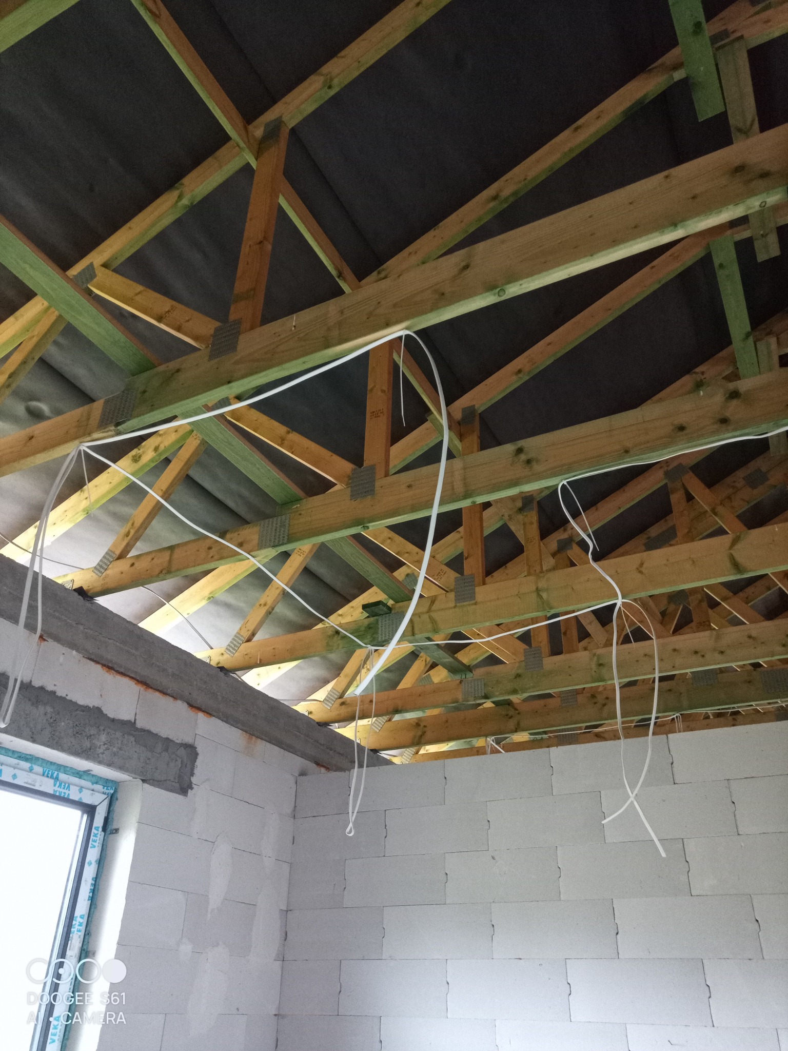 Surowy stan poddasza z widoczną drewnianą konstrukcją dachu i prowizorycznie poprowadzonymi przewodami elektrycznymi, widok z dołu na tle szarej membrany dachowej.