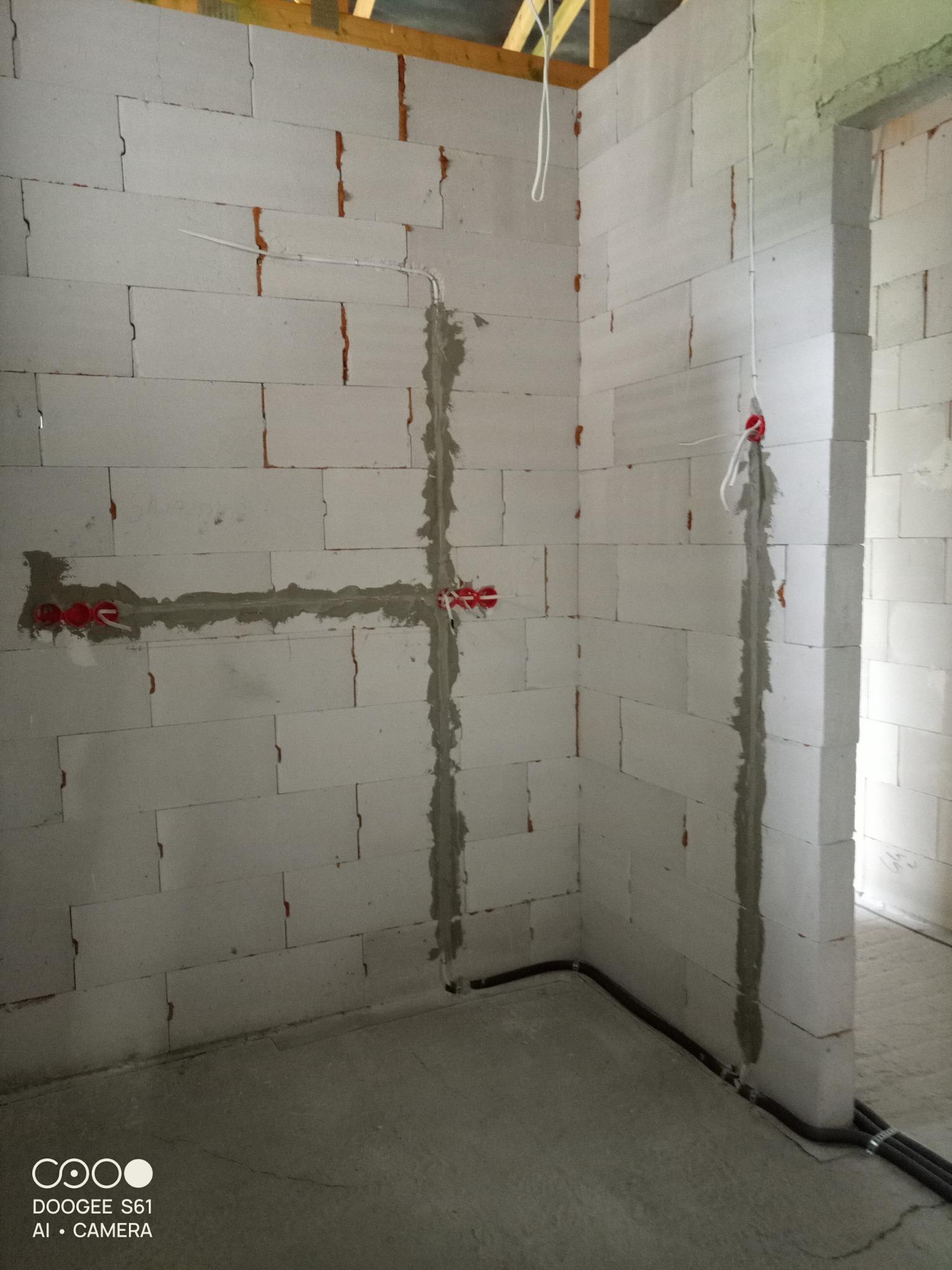 Wnętrze w trakcie prac instalacyjnych: przewody elektryczne prowadzone w bruzdach w ścianie z betonu komórkowego, zabezpieczone gipsem, widoczne puszki instalacyjne z czerwonymi elementami...