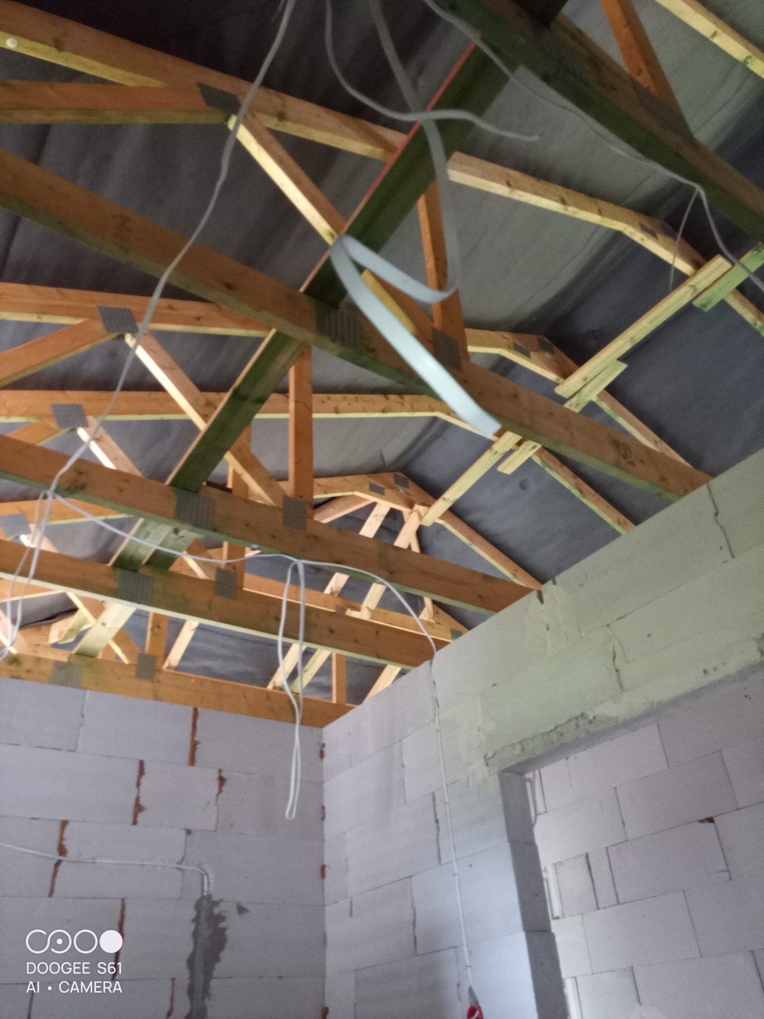 Wnętrze w trakcie budowy z widoczną drewnianą konstrukcją dachu i przewodami elektrycznymi zwisającymi z sufitu, częściowo poprowadzonymi po ścianach z białych bloczków.