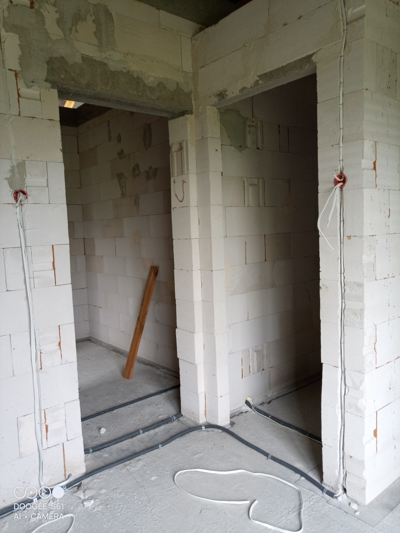 Surowy stan budynku z białych bloczków, widoczne dwa otwory drzwiowe, wystające kable elektryczne w peszlach na podłodze i w ścianach, instalacja elektryczna w trakcie montażu.