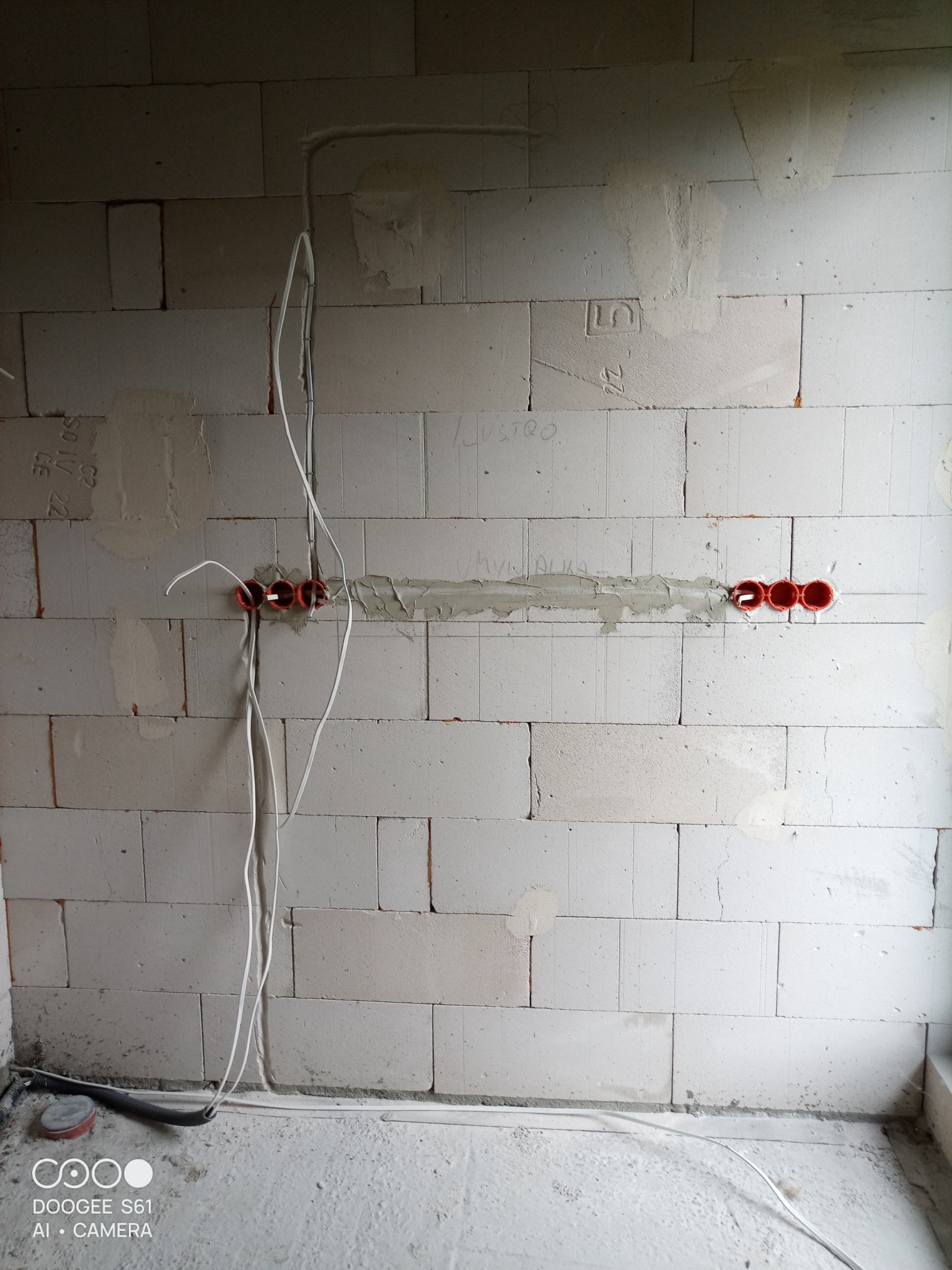 Surowa ściana z betonu komórkowego z trzema rzędami czerwonych puszek elektrycznych, z których wystają białe kable. Ściana częściowo otynkowana, widoczne napisy. Na podłodze leżą czarne rurki...