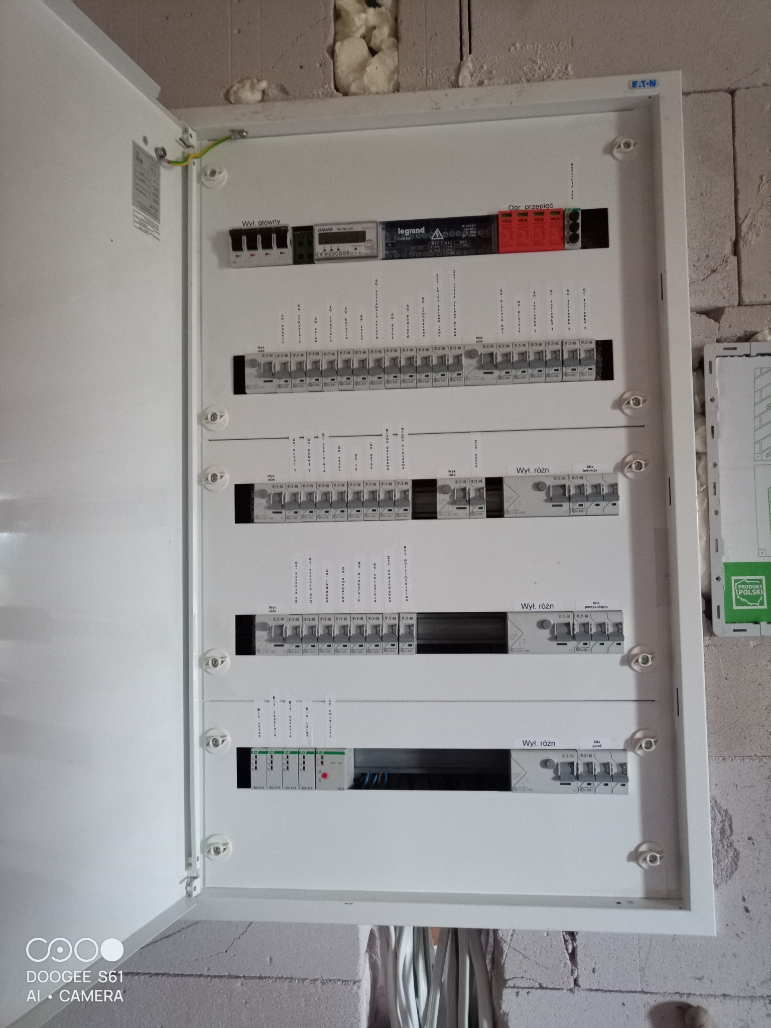Otwarta, biała skrzynka rozdzielcza z zainstalowanymi bezpiecznikami i wyłącznikami różnicowoprądowymi Eaton, Legrand i Orno na tle surowej ściany z betonu komórkowego i izolacji.