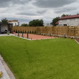 Nowoczesny ogród z równo przystrzyżonym trawnikiem, drewnianym tarasem, ozdobnym ogrodzeniem z bambusa i nasadzeniami z tuj i traw ozdobnych, oświetlony niskimi lampami ogrodowymi.