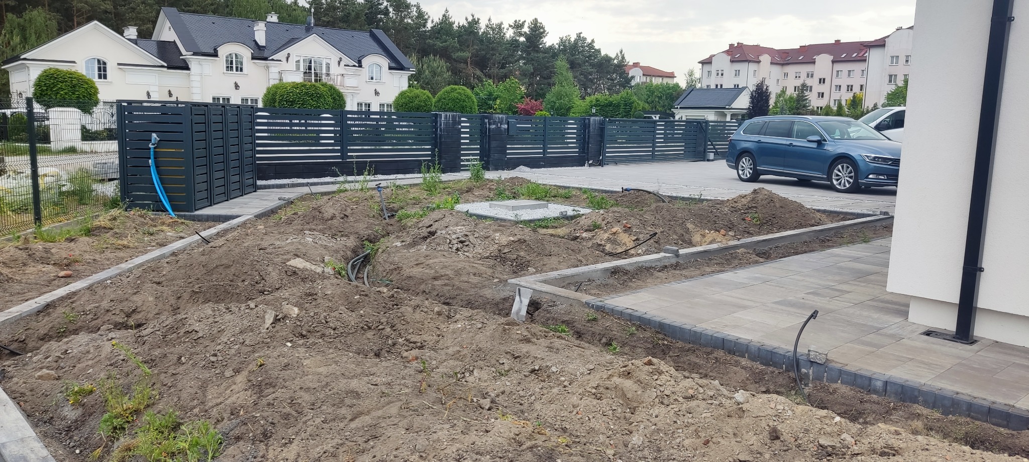 Przygotowanie terenu pod nowy ogród z widocznym ogrodzeniem panelowym, wykopanymi rowami na instalacje i częściowo ułożoną kostką brukową. Na dalszym planie domy i zaparkowany samochód.