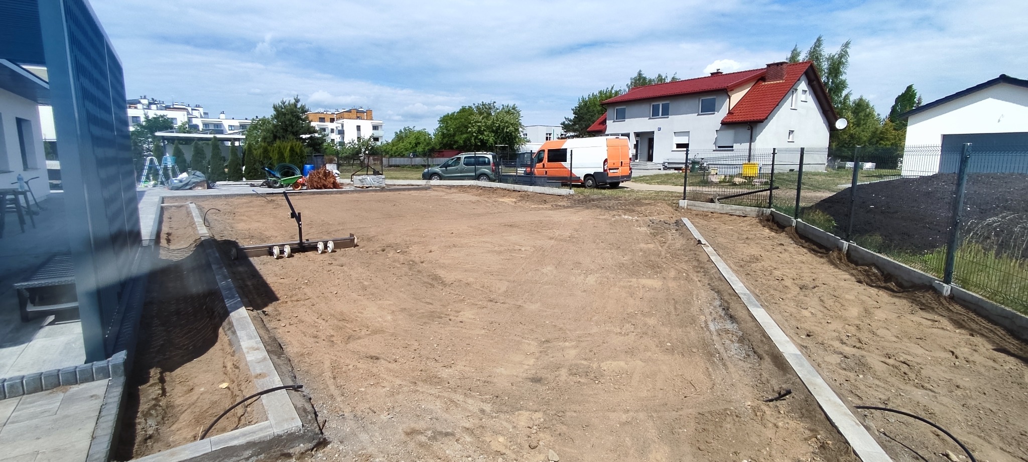 Przygotowany teren pod przyszły ogród z widocznymi obrzeżami trawnikowymi, wyrównaną ziemią, sprzętem do wyrównywania i dwoma samochodami dostawczymi w tle.