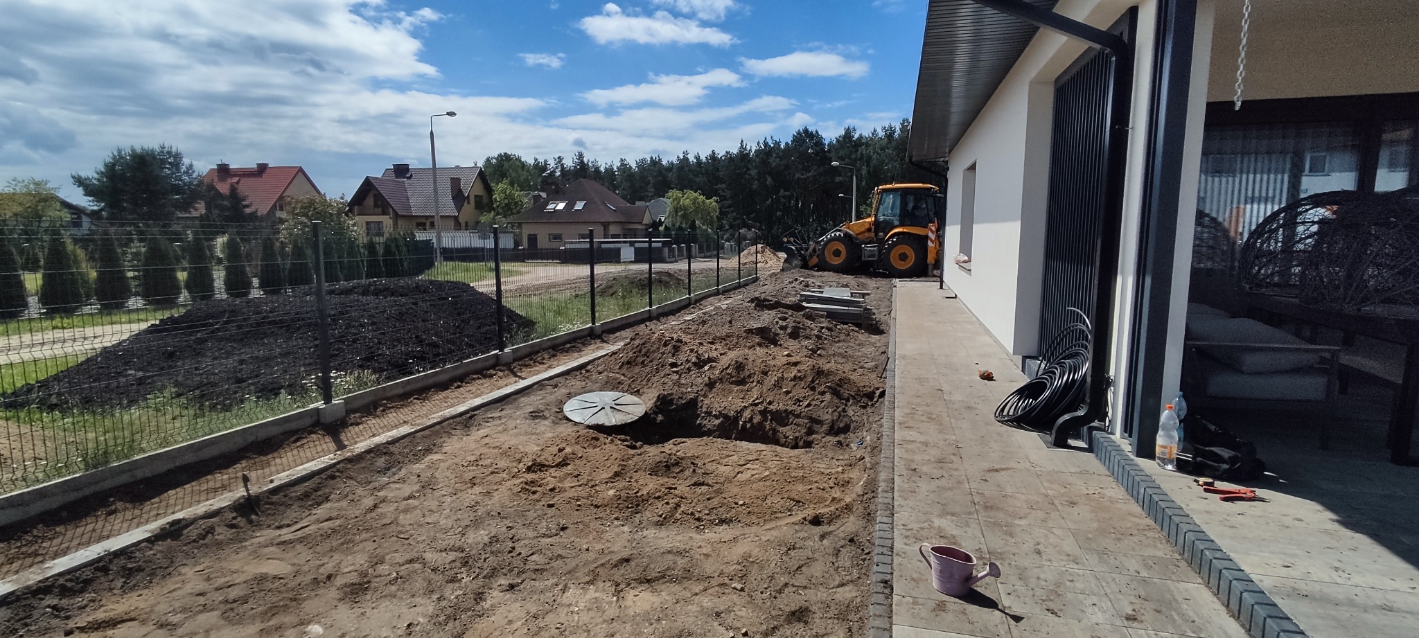 Żółta koparko-ładowarka na działce w trakcie prac ziemnych, widoczny wykop, nowa siatka ogrodzeniowa i przygotowane podłoże, w tle domy jednorodzinne.