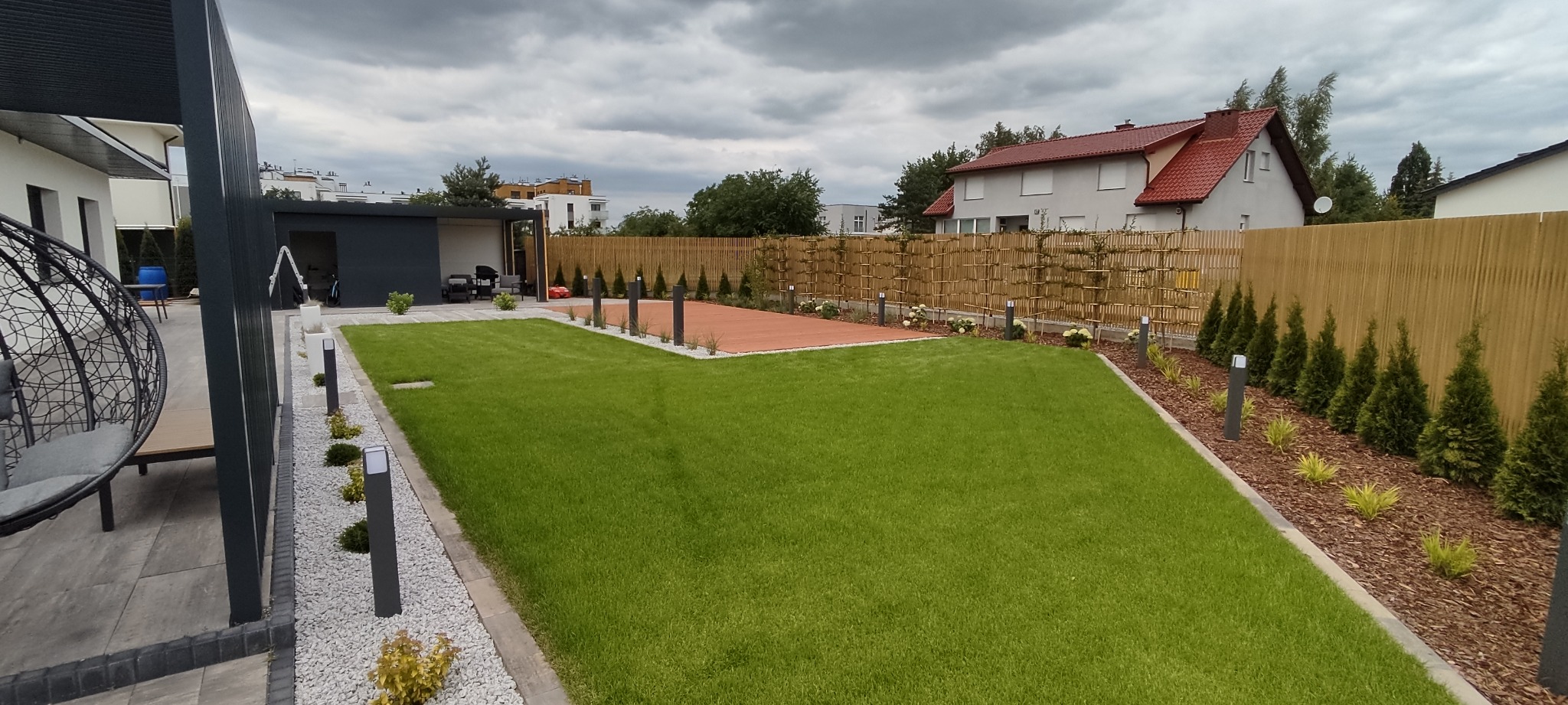 Nowoczesny ogród z równo przystrzyżonym trawnikiem, drewnianym tarasem, ozdobnym ogrodzeniem z bambusa i nasadzeniami z tuj i traw ozdobnych, oświetlony niskimi lampami ogrodowymi.