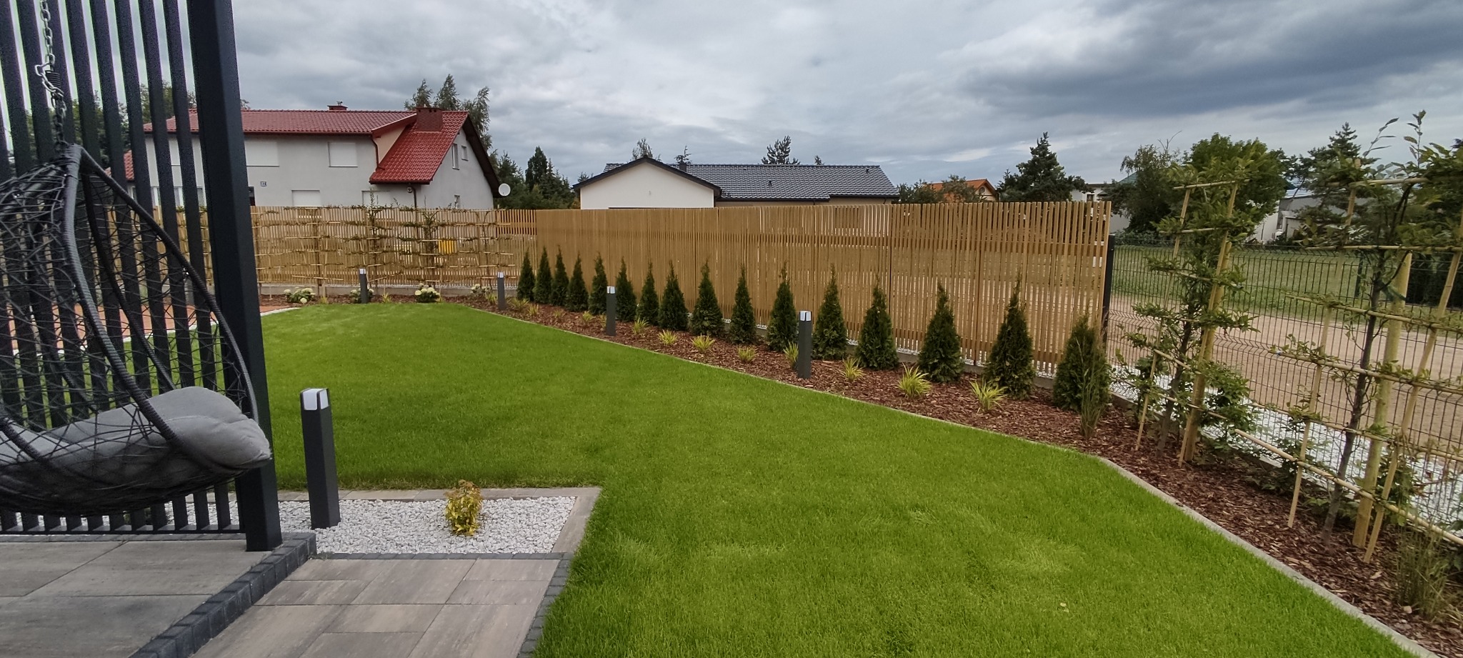 Nowoczesny ogród z równo przystrzyżonym trawnikiem, drewnianym ogrodzeniem z pionowych listew, rzędem małych tujek i lampkami solarnymi wzdłuż ogrodzenia, widok z tarasu z wiszącym fotelem.