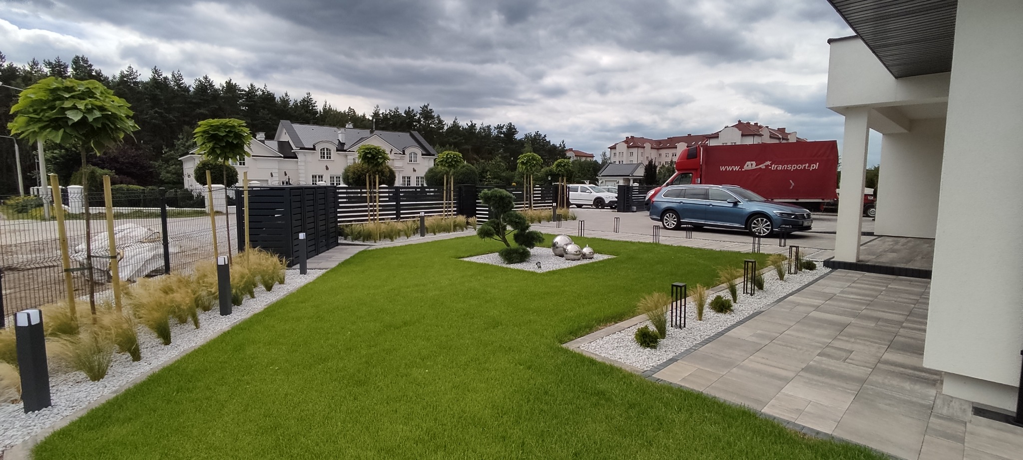 Nowoczesny ogród z równo przystrzyżonym trawnikiem, ozdobnymi krzewami, kulami ze stali nierdzewnej i oświetleniem wzdłuż ścieżki, na tle domu i drzew.