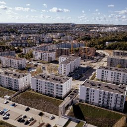 Szymon Napiwodzki - Widok z lotu ptaka na osiedle nowoczesnych, białych budynków mieszkalnych z balkonami, z parkingiem pełnym samochodów, otoczone zielenią i zabudową miejską w słoneczny dzień.