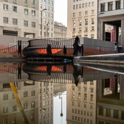 Szymon Napiwodzki - Architektura miejska odbita w kałuży: budynki, ażurowa konstrukcja i sylwetki przechodniów.