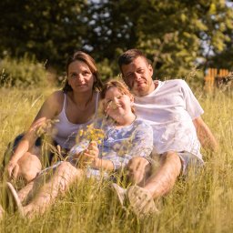 Szymon Napiwodzki - Rodzinne zdjęcie: mama, tata i córka siedzą na łące w wysokiej trawie, dziewczynka trzyma bukiecik żółtych kwiatów, w tle drzewa i dom z czerwonym dachem.