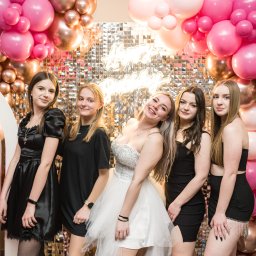 Szymon Napiwodzki - Grupa pięciu młodych kobiet w sukienkach pozuje na tle błyszczącej ściany i dekoracji z różowych i złotych balonów.