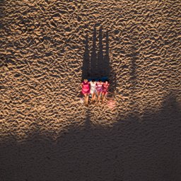 Szymon Napiwodzki - Widok z góry na cztery kobiety leżące na kocu na piaszczystej plaży, rzucające długie cienie w popołudniowym słońcu.