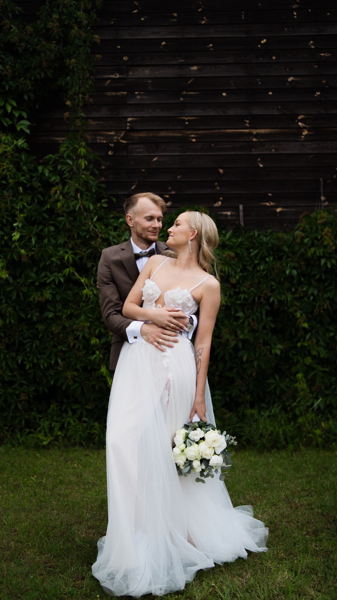 Uśmiechnięta para młoda w objęciach na tle drewnianej ściany porośniętej bluszczem, panna młoda trzyma bukiet białych róż.